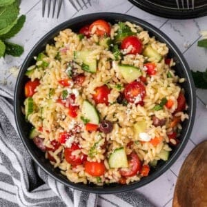 A bowl of orzo pasta salad with cherry tomatoes, cucumber slices, black olives, red onions, and crumbled feta cheese. The pasta salad is garnished with fresh parsley and placed on a white marble surface next to a striped cloth and utensils.
