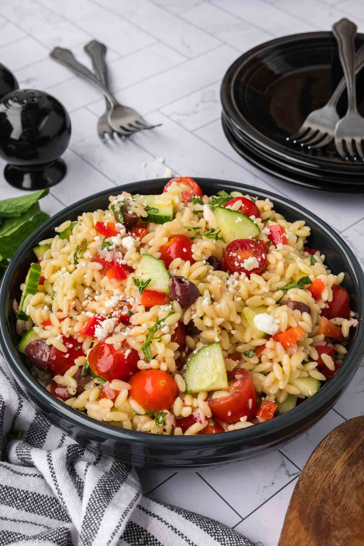 A bowl of orzo salad filled with fresh vegetables like cherry tomatoes, cucumber, and red onions, topped with crumbled feta cheese and olives. The dish is placed on a white tiled surface next to a black pepper grinder, a striped napkin, and a stack of black plates with forks.