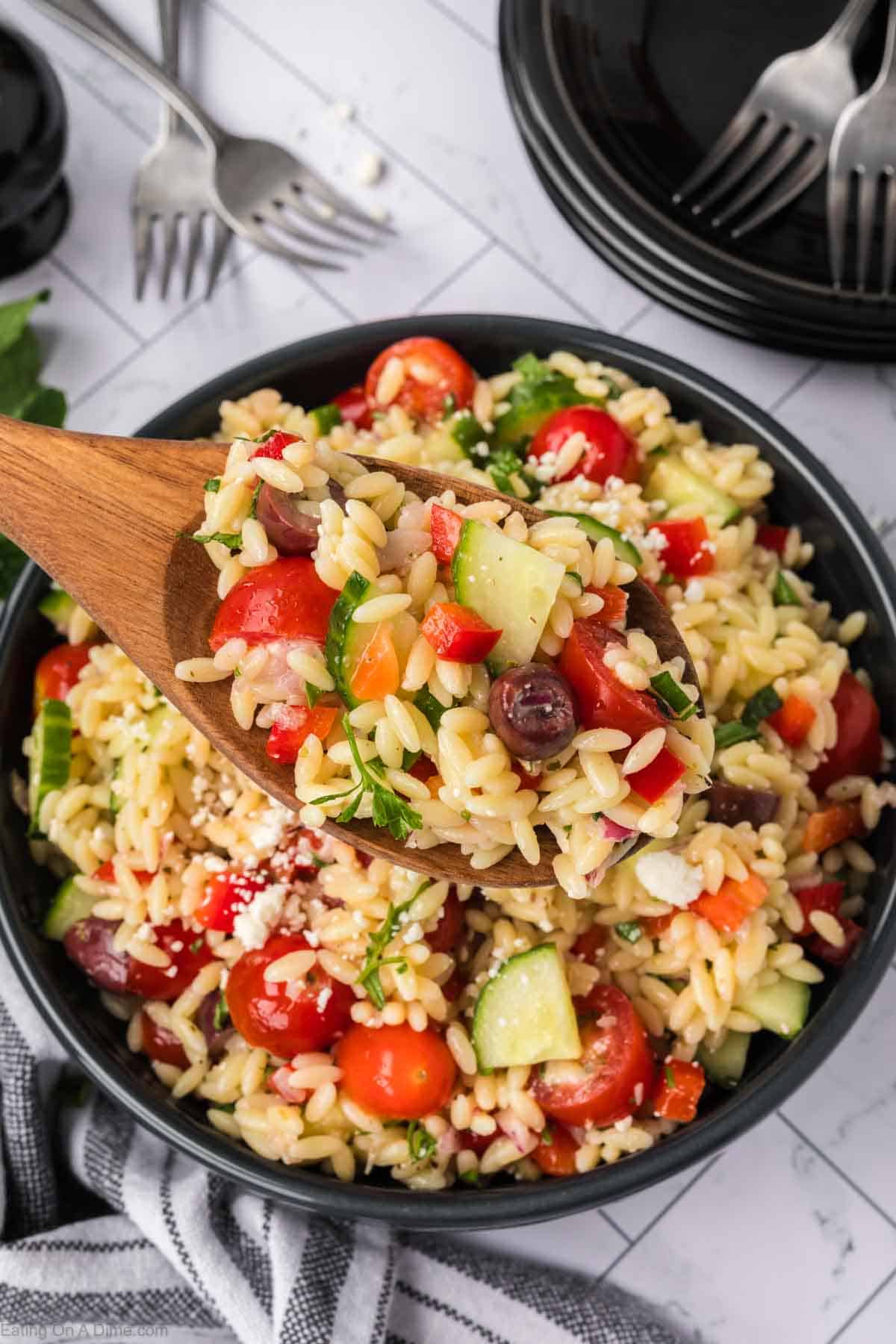 A wooden spoon lifting a serving of colorful orzo pasta salad from a black bowl. The vibrant salad, featuring orzo pasta, cherry tomatoes, cucumbers, olives, bell peppers, feta cheese, and fresh herbs, is full of flavor. In the background, there are forks, plates, and a striped napkin.