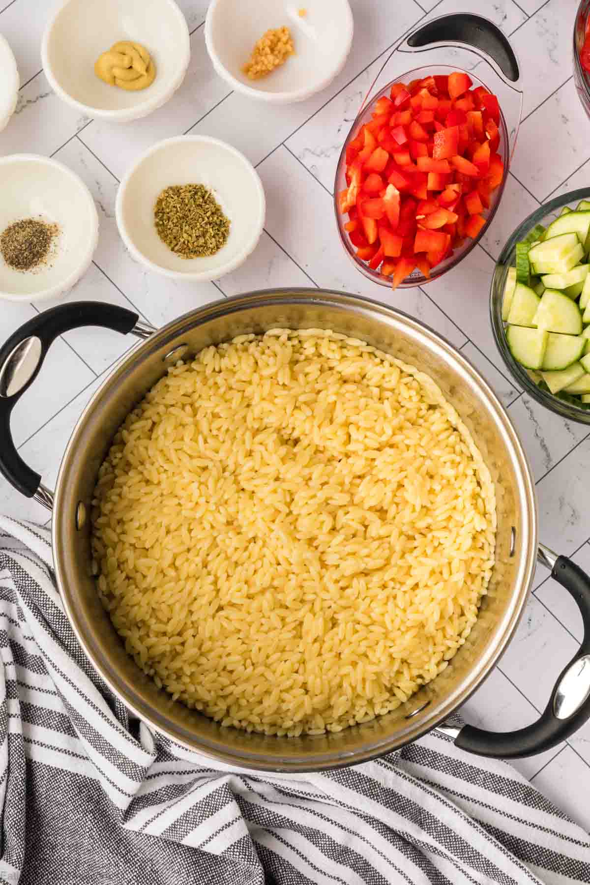 A pot filled with cooked orzo pasta is centered on a countertop. Surrounding it are small white bowls containing seasonings and spices, and glass bowls with chopped red peppers and cucumber, ready for a fresh orzo pasta salad. A striped kitchen towel is partially visible to the left of the pot.