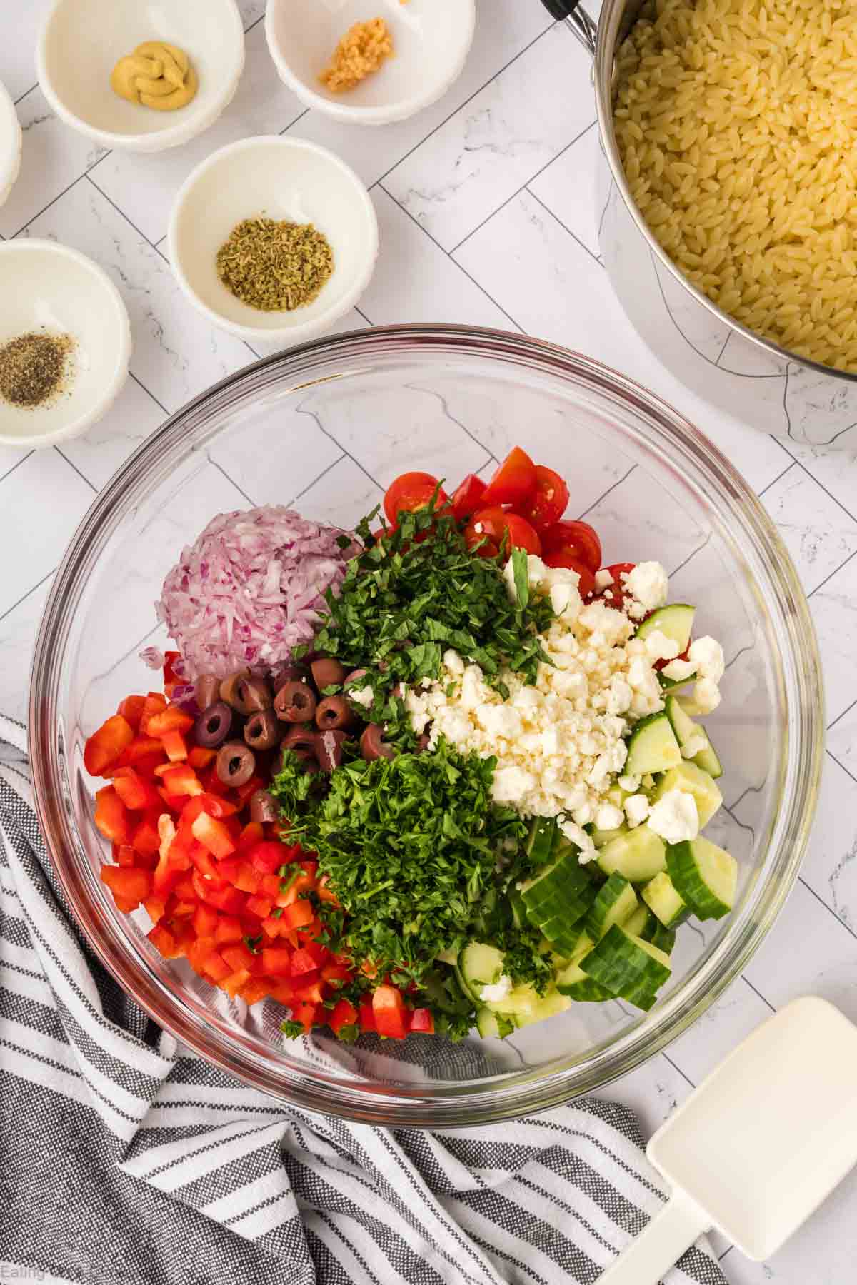 A glass bowl containing chopped red onions, red bell peppers, cherry tomatoes, black olives, cucumber, parsley, and feta cheese sits nearby a pot of cooked orzo pasta. Small bowls with seasonings and minced garlic rest on a white tiled surface with a striped towel, ready for a delicious orzo salad.