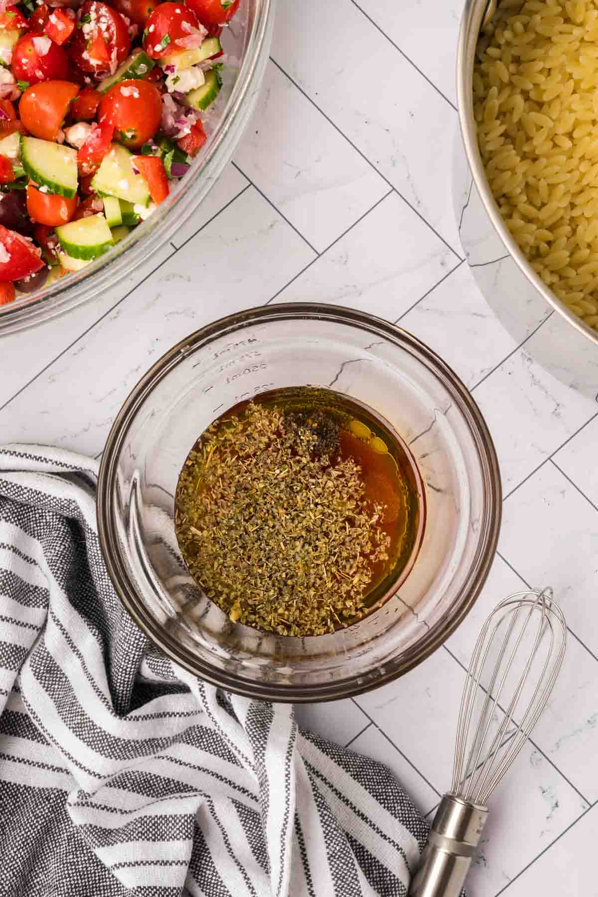 A glass bowl filled with herbs and oil sits on a white tiled surface next to a metal pot of orzo pasta, a striped kitchen towel, a whisk, and a bowl of chopped cucumber and tomato salad. The ingredients are ready for mixing.