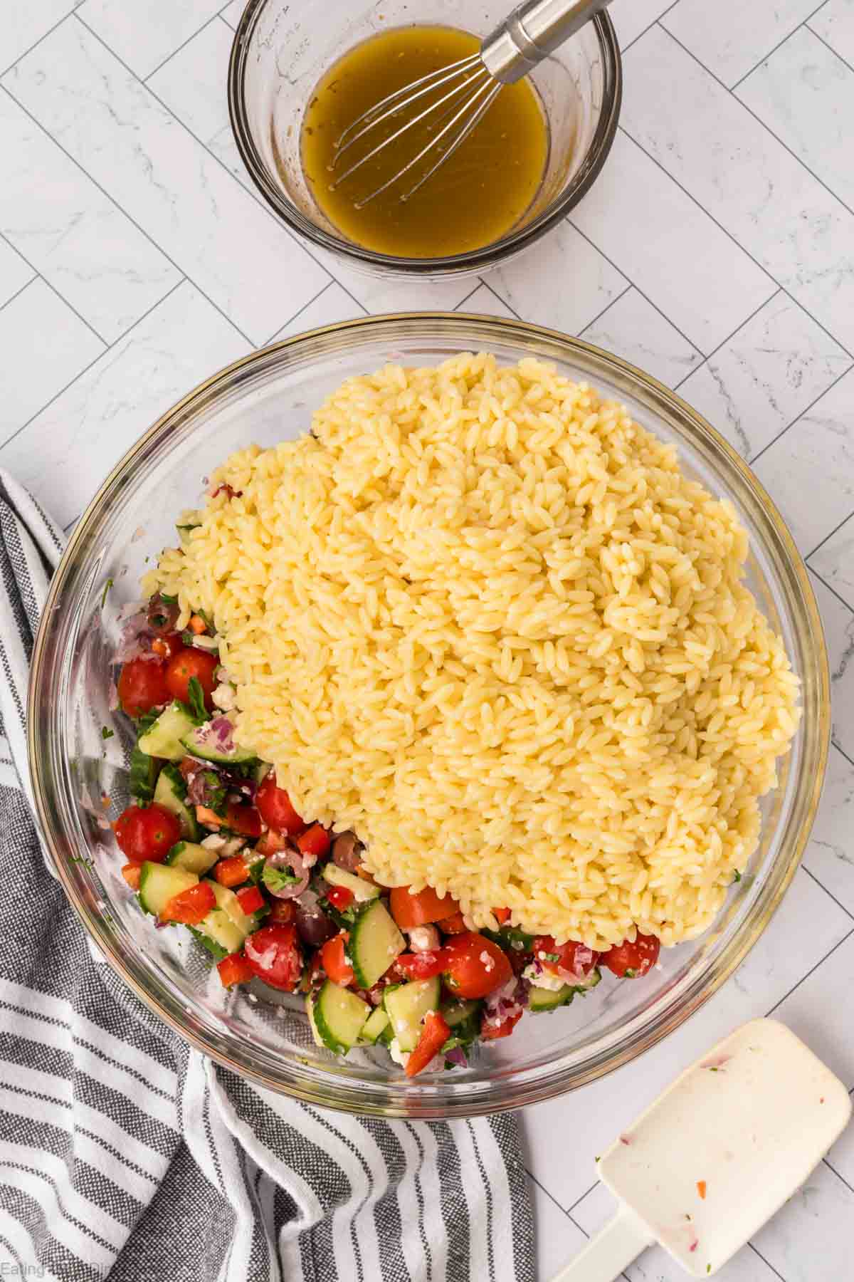 A large glass bowl filled with a partially mixed orzo salad, containing chopped cucumbers, tomatoes, red onions, and feta cheese. A small bowl with whisked dressing sits nearby, along with a white spatula placed on a striped cloth.