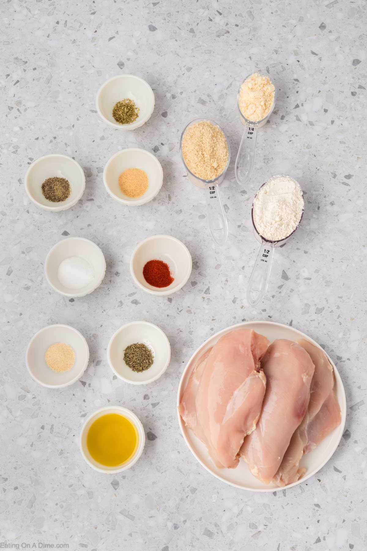 A top-down view of a light gray countertop showcases raw chicken breasts on a white plate, surrounded by small bowls of various spices, and measuring spoons filled with panko breadcrumbs and flour. A small bowl of oil is also present, perfect for preparing Shake and Bake Chicken.