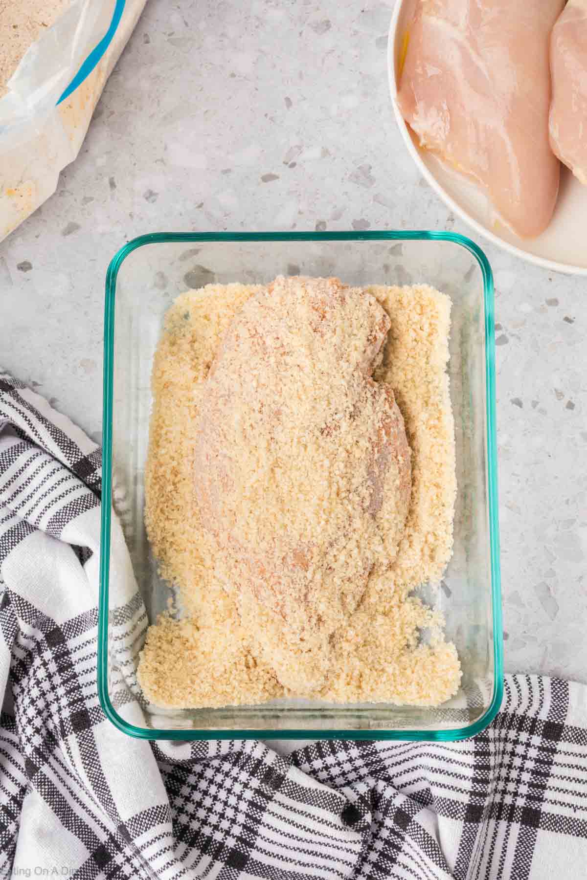 A Shake and Bake breaded chicken breast sits in a glass container filled with breadcrumbs on a gray countertop. Nearby, there is a white bowl containing raw chicken breasts and a black and white checkered cloth partially visible.