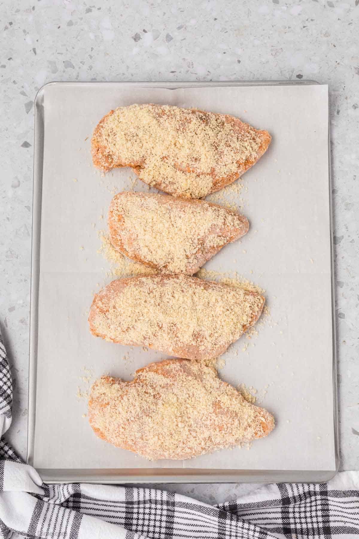 Four Shake and Bake Chicken breasts placed on a parchment-lined baking sheet. A black and white checkered kitchen towel is partially visible in the lower left corner of the image. The background is a terrazzo-style countertop.