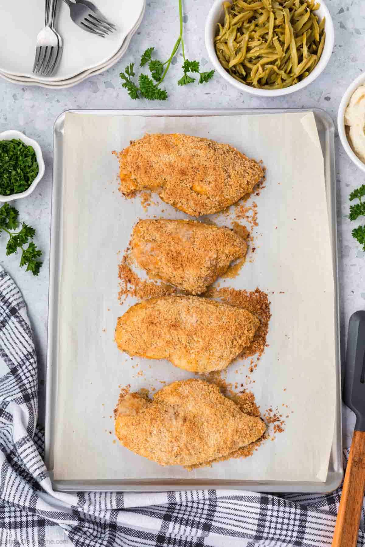 A baking sheet topped with four Shake and Bake Chicken breasts aligned in two rows. Surrounding the sheet are a cloth napkin, a spatula, a bowl of green beans, a dish of mashed potatoes, and a small bowl of chopped parsley. Plates and forks are visible in the background.