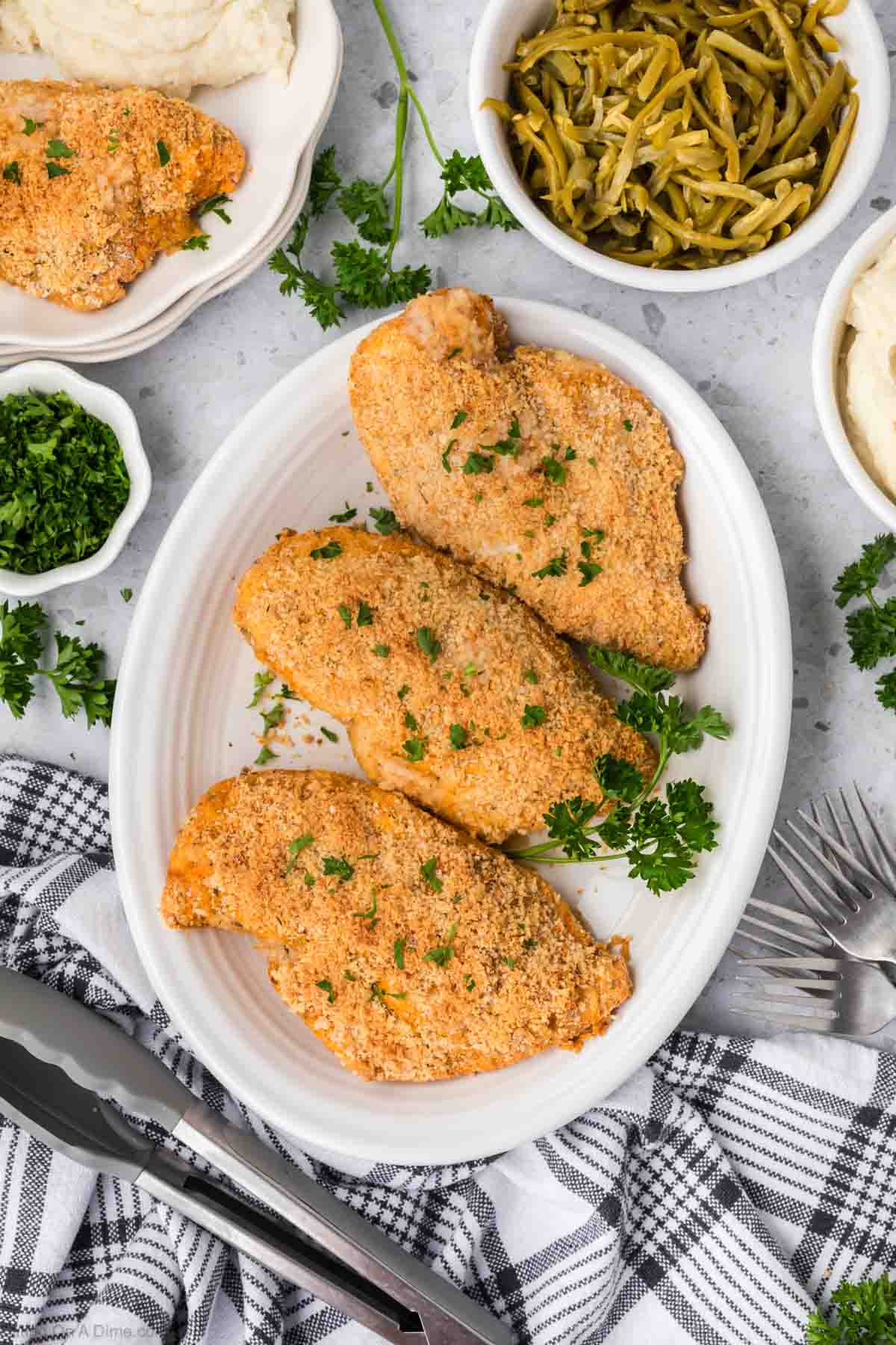 A white plate with three Shake and Bake Chicken breasts garnished with parsley. Surrounding the plate are bowls of mashed potatoes, green beans, and chopped parsley. The table setting includes utensils and a checkered cloth napkin.