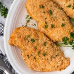 A white plate holds two Shake and Bake chicken breasts garnished with chopped parsley. The chicken is golden brown, crispy on the outside, and appears to be seasoned with herbs. A parsley sprig is placed on the side for decoration, completing this delightful recipe.
