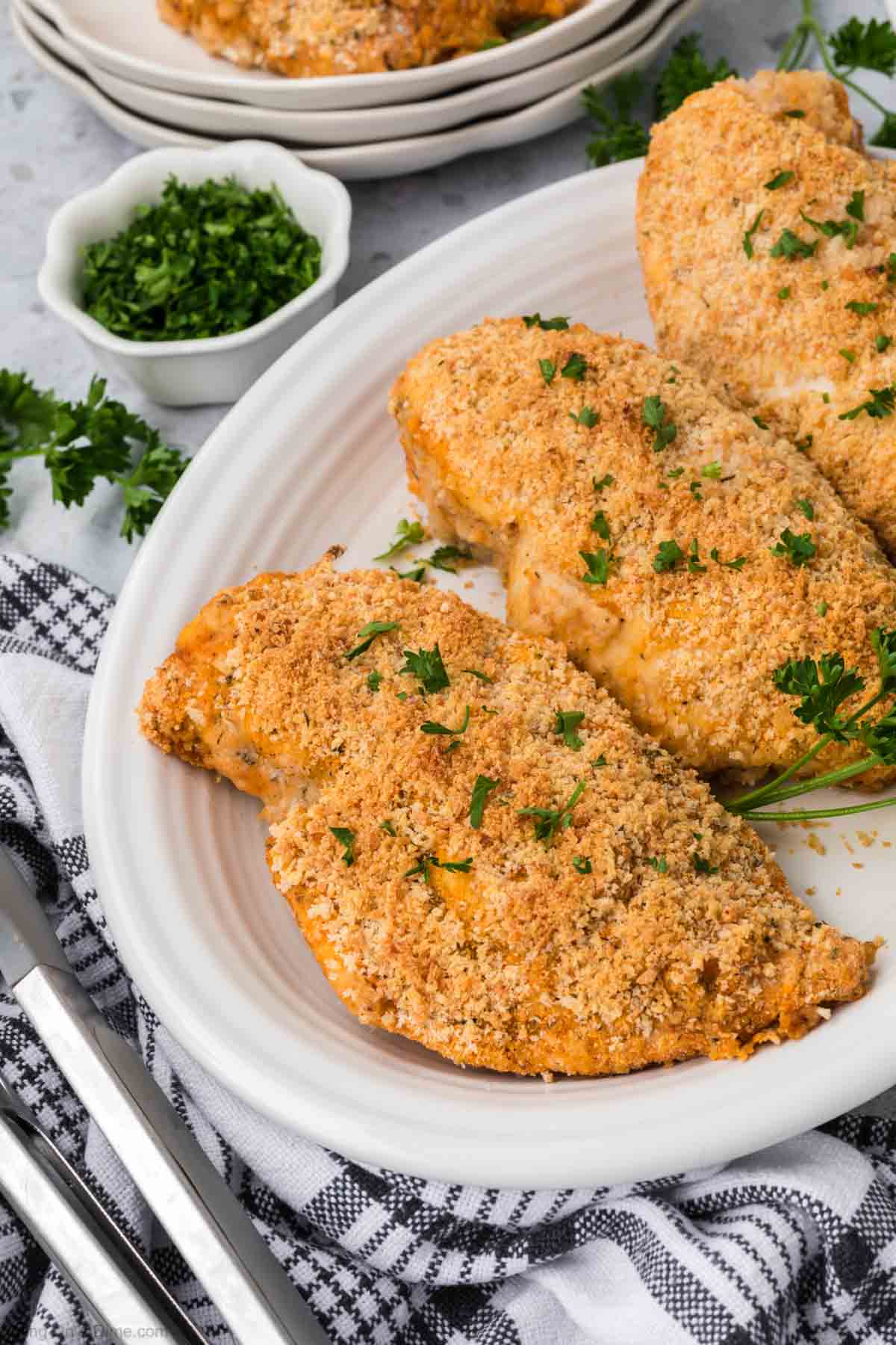 A white plate holding three pieces of Shake and Bake chicken, garnished with chopped parsley. Beside the plate, there is a small bowl filled with more chopped parsley. A black and white checkered cloth and metal utensils are partially visible in the foreground.