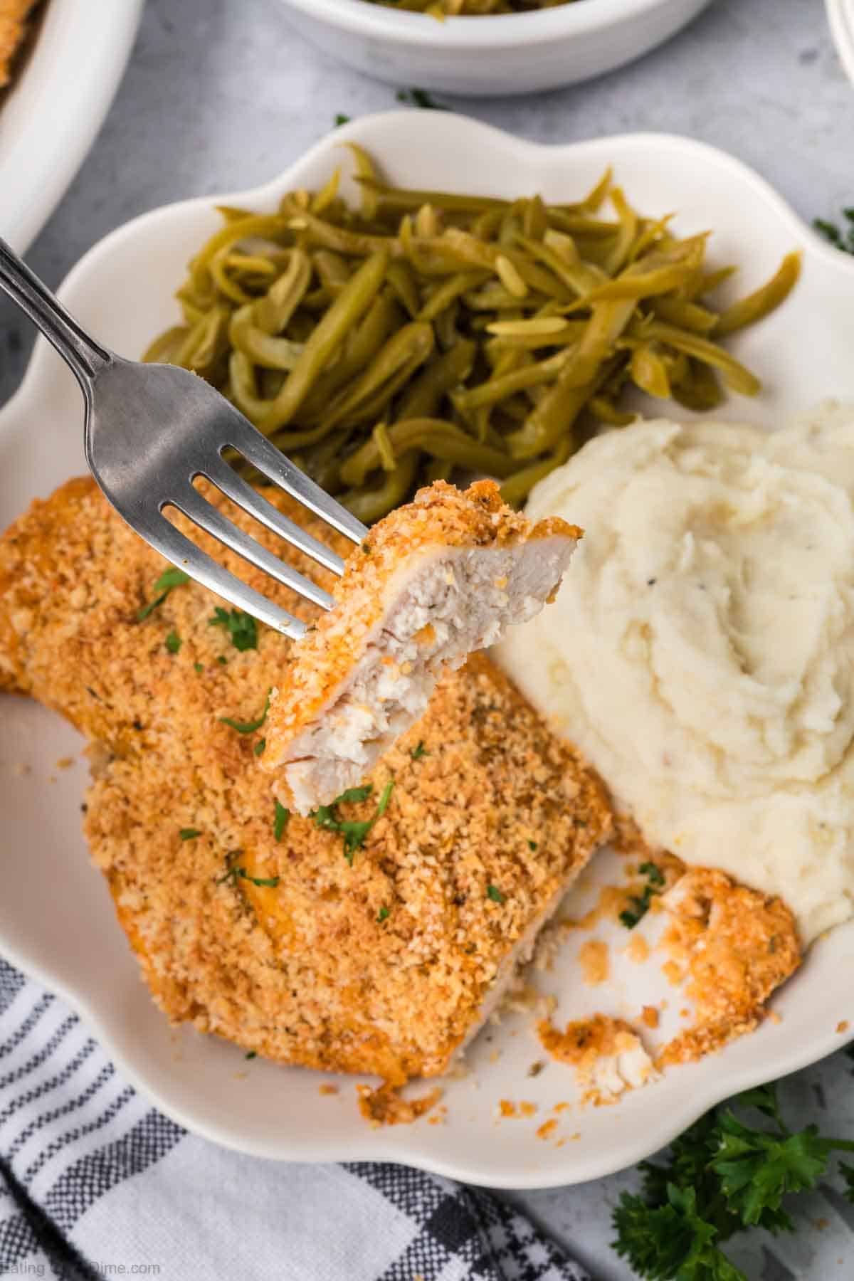 A fork holds a bite of Shake and Bake Chicken breast with a serving of green beans and mashed potatoes on a white plate. The scene suggests a comforting, homemade meal. The table is set with a striped cloth and a garnish of parsley is visible.