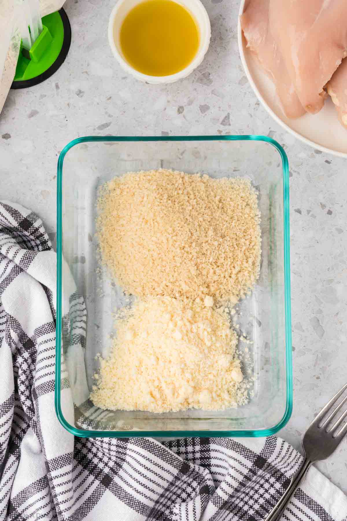 A glass container filled with breadcrumbs and grated cheese sits on the counter next to a small bowl of oil, raw chicken breasts on a plate, and a black-and-white checkered towel. A fork is also visible nearby. It's all set up for tonight's Shake and Bake Chicken.