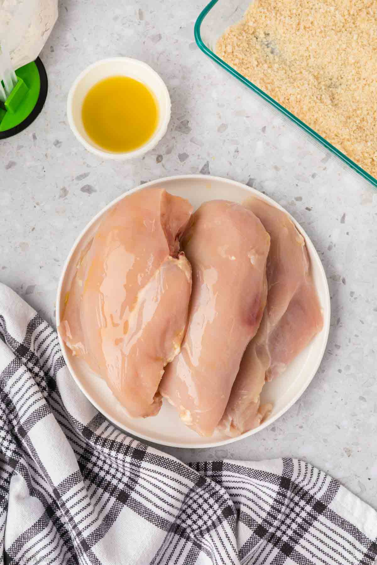 A plate with raw chicken breasts sits on a countertop next to a white bowl of olive oil and a baking dish containing bread crumbs, ready for the Shake and Bake Chicken. A black and white checkered cloth is draped nearby, against a backdrop of light-colored marble.