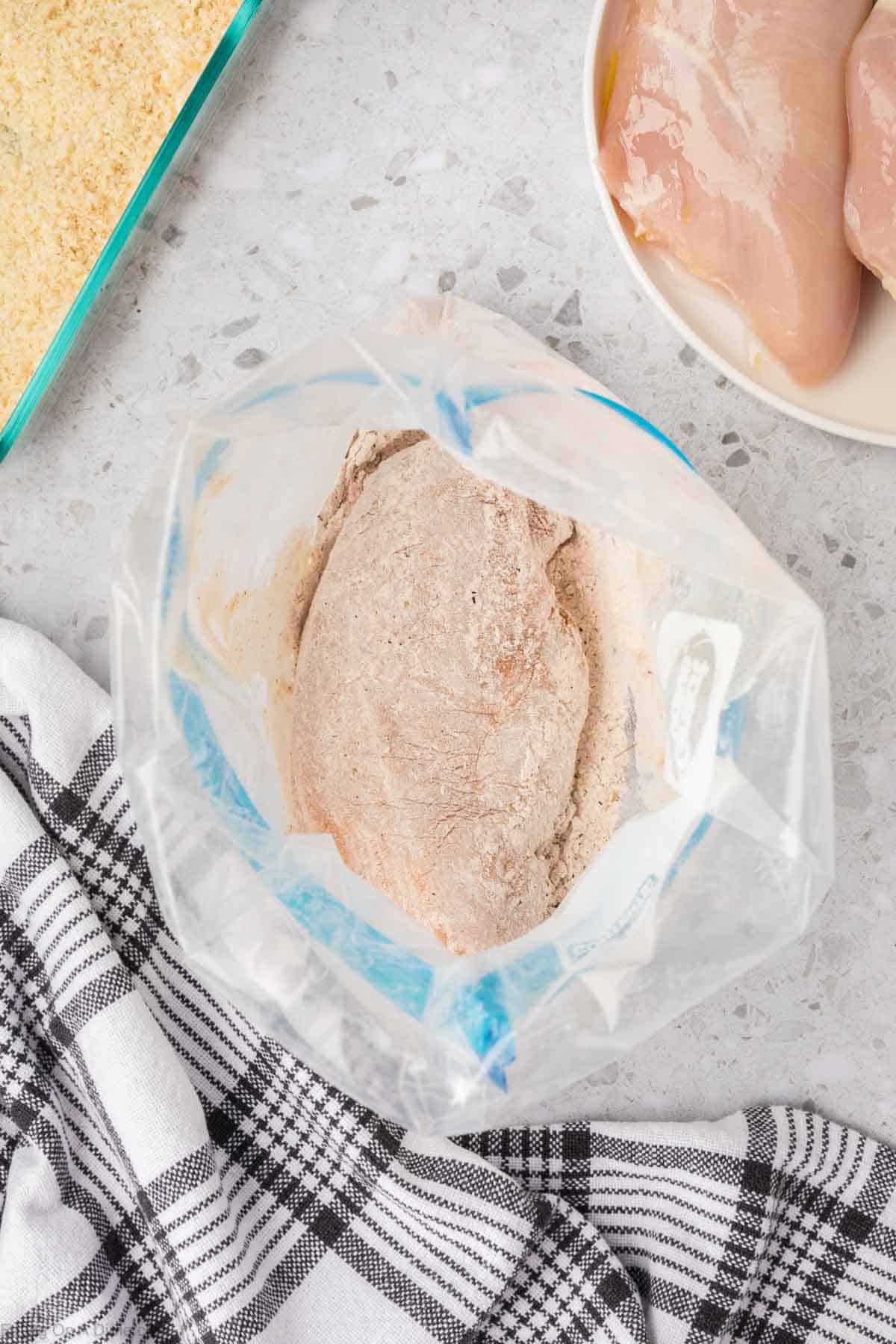 A raw chicken breast in a plastic bag filled with flour and seasonings, ready for coating. Nearby, there is a plate with additional raw chicken breasts and a dish with breadcrumbs for your Shake and Bake Chicken recipe. A black and white checkered cloth is placed next to the bag on the countertop.