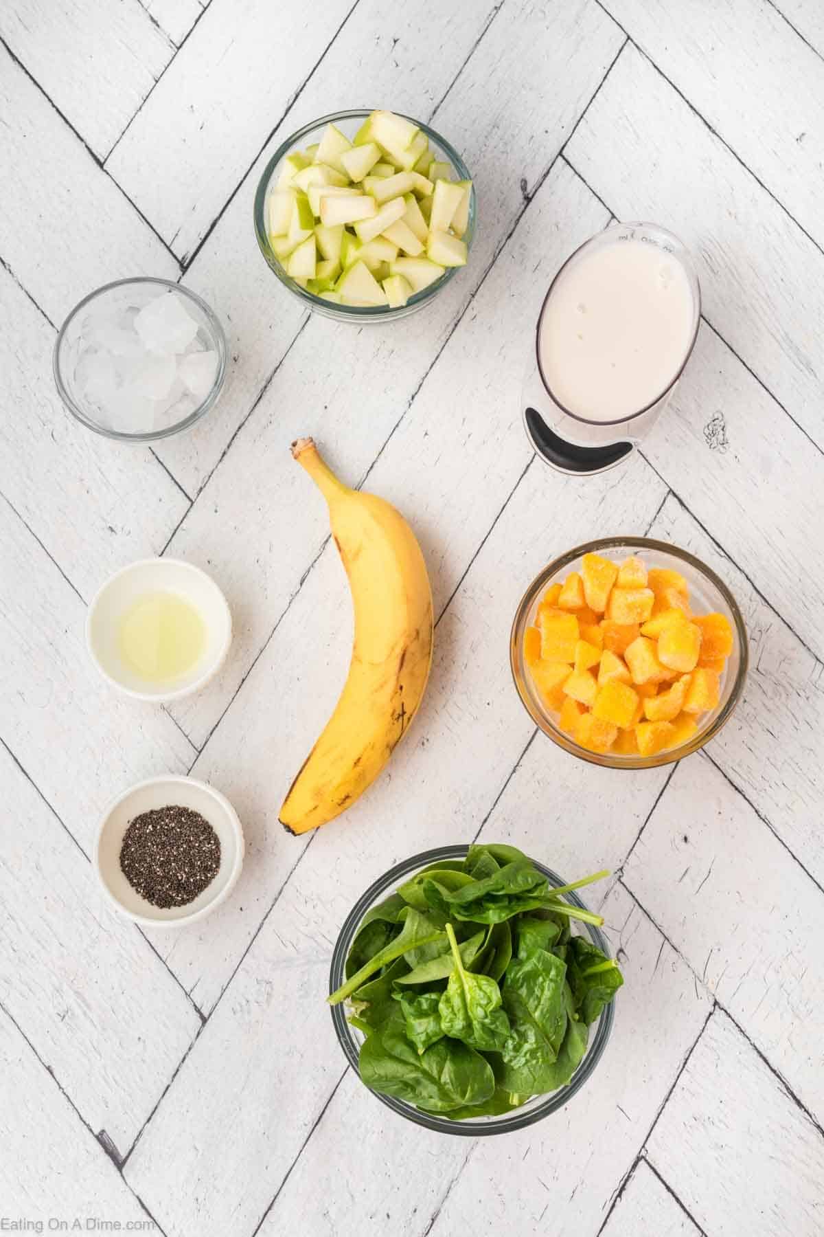 Overhead view of smoothie ingredients: a bowl of chopped green apples, a bowl of ice, a glass of milk, a bowl of cubed mango, a bowl of spinach, a whole banana, a small bowl of chia seeds, and lemon juice—all arranged on a white herringbone-patterned surface. Perfect for crafting your ultimate fruit smoothie.