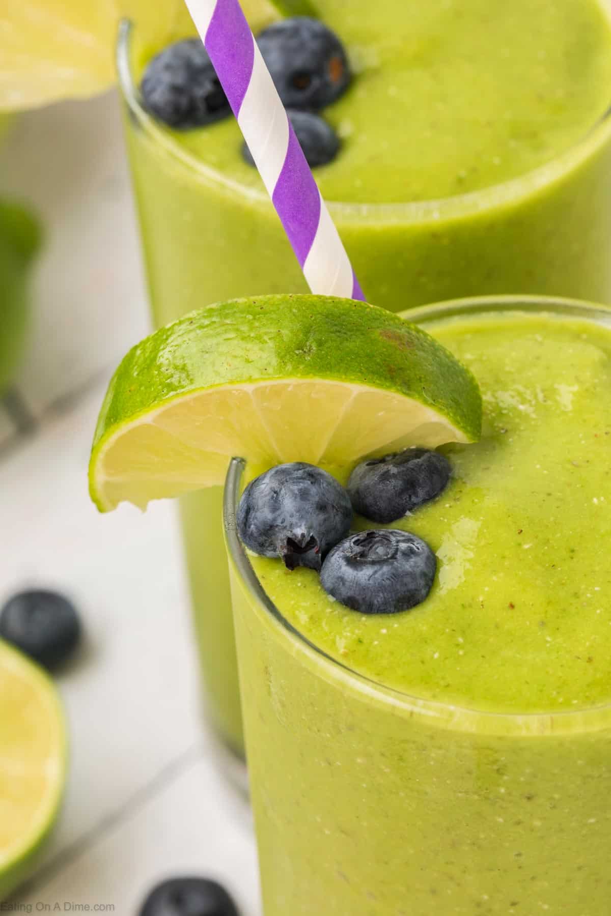 A close-up of a green smoothie, enriched with spinach, topped with a slice of lime and several blueberries. A purple and white striped straw is inserted into the vibrant drink, served in a clear glass, with additional blueberries and lime slices visible in the background.