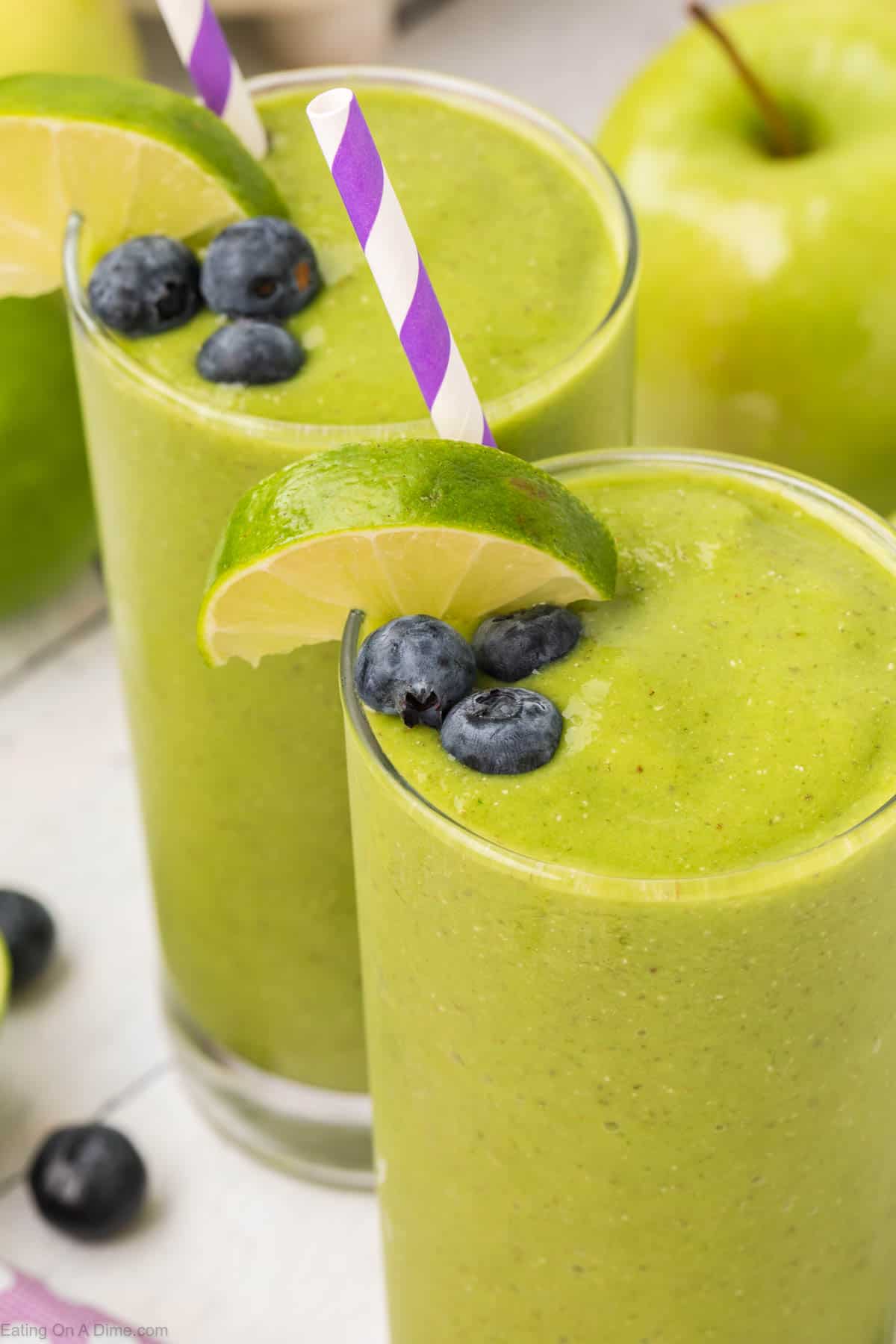 Two tall glasses of green smoothie, enriched with spinach, topped with fresh blueberries and a wedge of lime, each adorned with a purple and white striped straw. A whole green apple and more blueberries are visible in the background. The fruit smoothies appear thick and creamy.