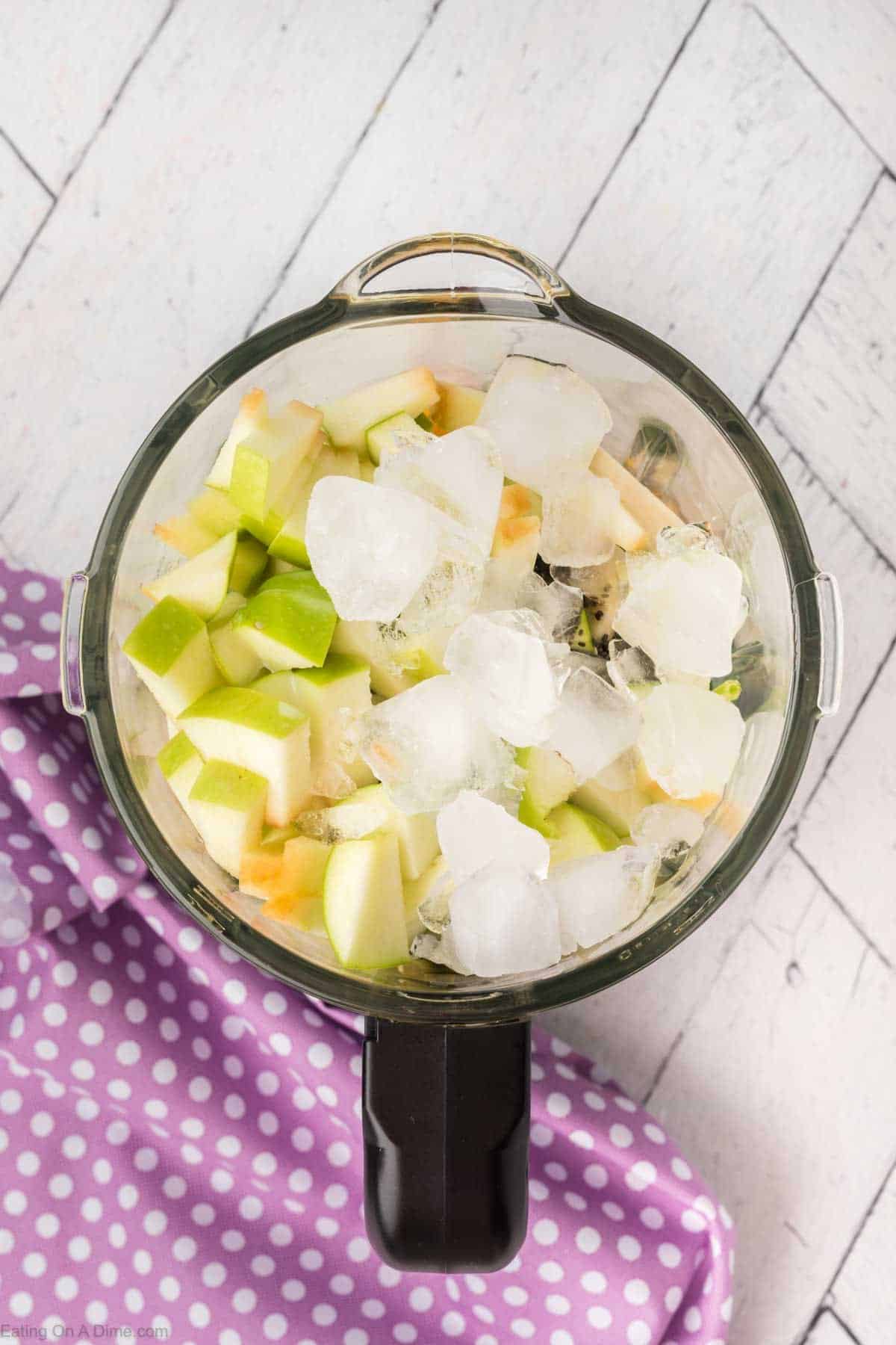 A blender pitcher filled with chopped green and white apple pieces, spinach, and ice cubes sits on a white wooden surface. A purple cloth with white polka dots is partially visible underneath the blender, ready for a nutritious smoothie.