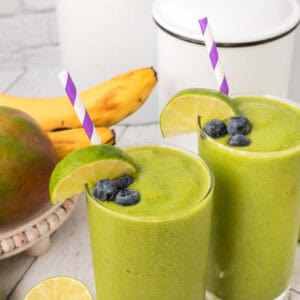 Two glasses of green smoothie topped with blueberries, lime slices, and purple-striped straws are placed on a white surface. A whole mango, bananas, a lime slice, and a white canister are in the background. This vibrant fruit blend is perfect for those who love spinach in their smoothie.