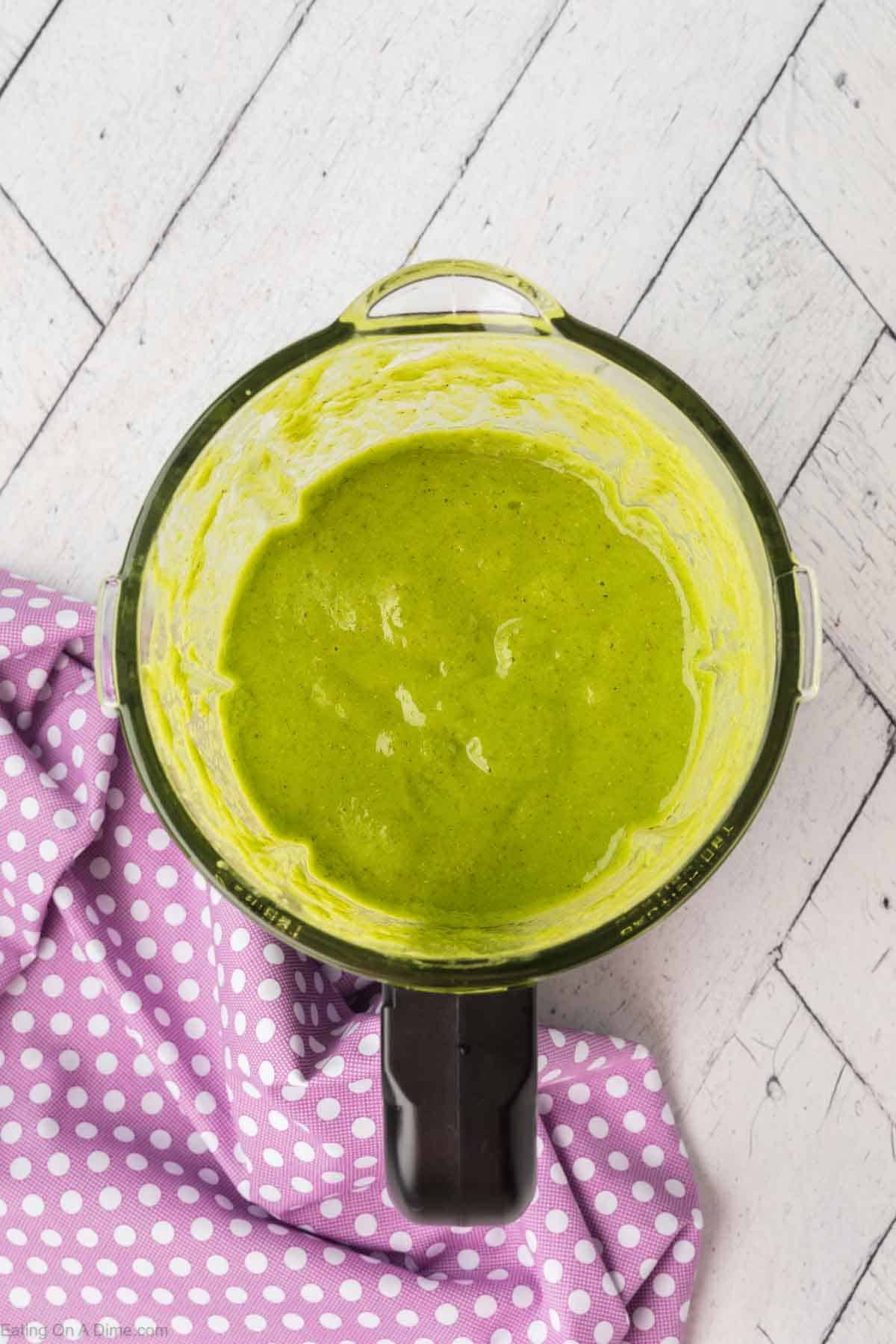 A blender filled with a smooth, green spinach smoothie sits on a white wooden surface. Next to the blender is a purple cloth with white polka dots, partially visible in the bottom left corner.