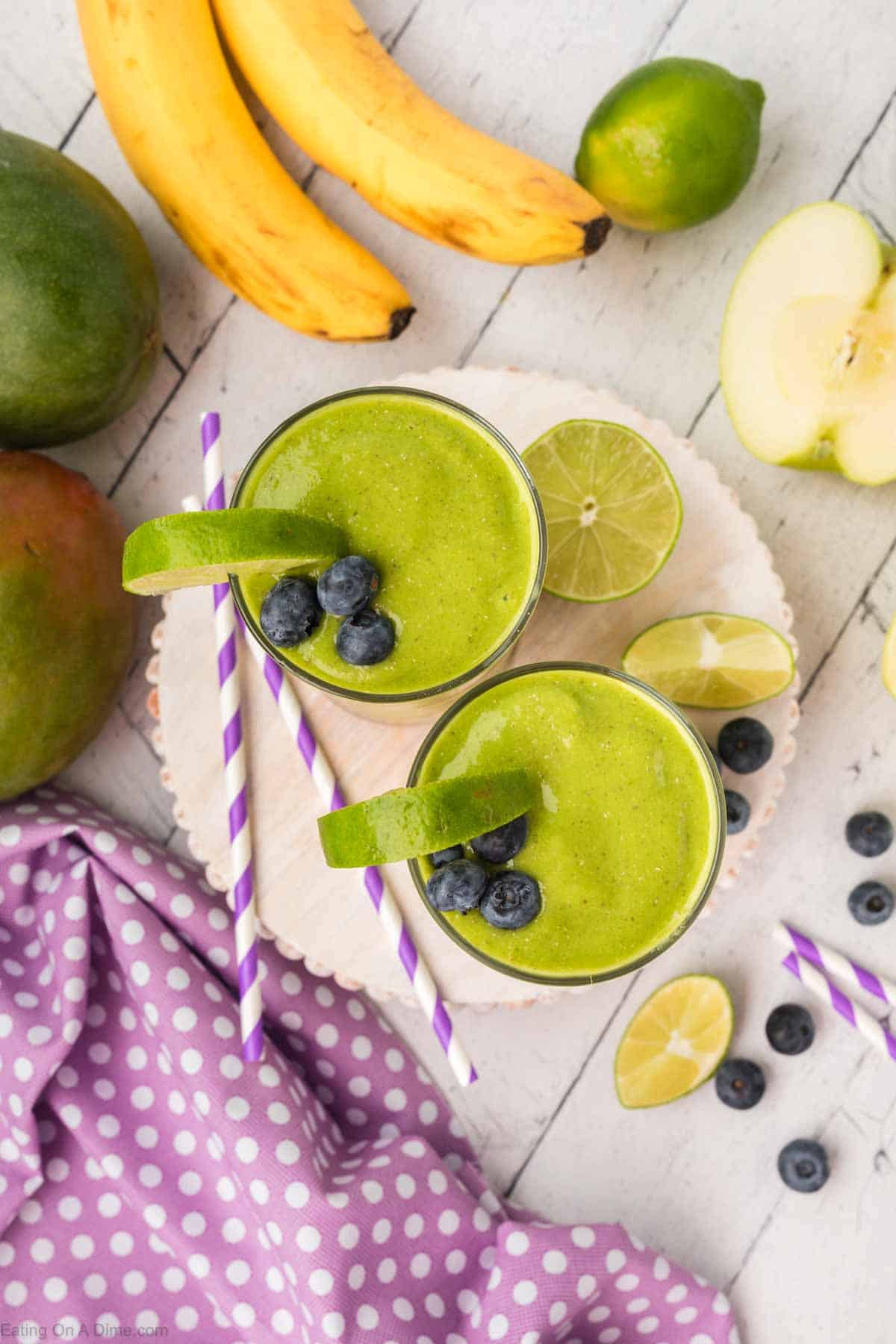 Two green smoothies, blended with fresh spinach, are garnished with lime wedges and blueberries, served with purple and white striped straws. Surrounding the smoothies are an array of vibrant fruit: bananas, a lime, a green apple, mangoes, plus extra lime wedges and blueberries. A purple polka dot cloth is visible nearby.