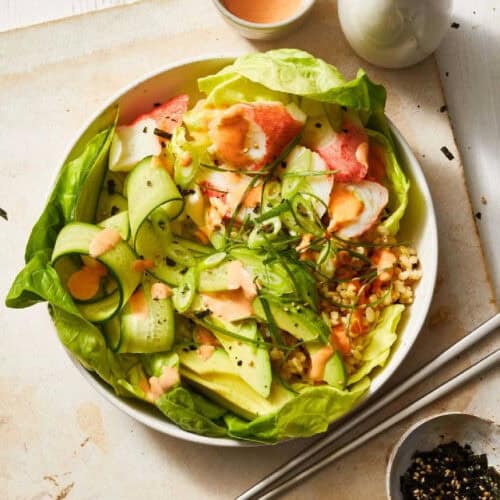 A bowl containing a fresh salad with lettuce, cucumber, avocado, and chunks of seafood, topped with a creamy dressing. The rice bowl is placed on a white surface with chopsticks, a small dish of seasoning, and a sauce bottle nearby.