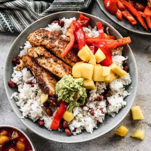 A delightful rice bowl featuring grilled chicken strips, diced mango, red bell peppers, black beans, and white rice, topped with a scoop of guacamole. A striped cloth and a small dish of salsa are visible in the background. Perfect for those seeking vibrant rice recipes.