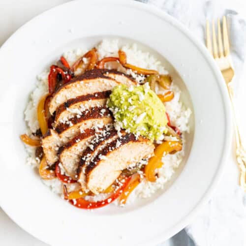 A white plate featuring a rice bowl recipe with sliced seasoned chicken breast on a bed of white rice, accompanied by sautéed bell peppers and onions. A dollop of guacamole and a sprinkle of crumbled cheese sit atop the dish. Gold cutlery and a light-colored cloth are placed beside the plate.