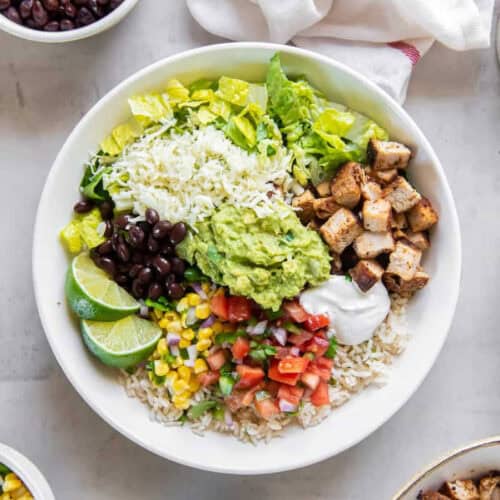 A bowl filled with chopped lettuce, shredded cheese, black beans, lime wedges, guacamole, grilled chicken chunks, sour cream, corn, diced tomatoes, and rice. The ingredients are arranged in sections to inspire rice bowl recipes that create a colorful and appetizing meal.