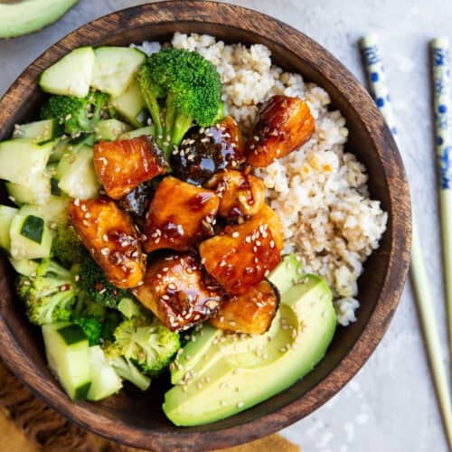 A wooden bowl filled with a healthy meal consisting of cubed teriyaki tofu topped with sesame seeds, sliced avocado, steamed broccoli, cucumber pieces, and brown rice. Perfect for those exploring rice bowl recipes. Chopsticks rest beside the bowl on a light surface.