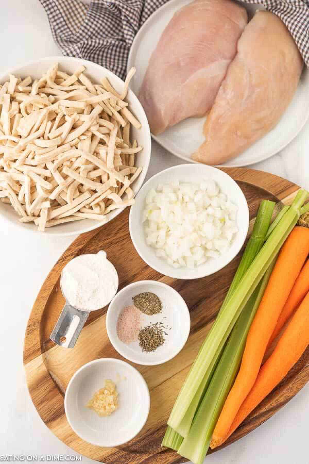 Close up image of ingredients for chicken noodle soup which consist of egg noodles, chicken breast, chopped onions, flour, garlic, seasoning, carrots, and celery. 