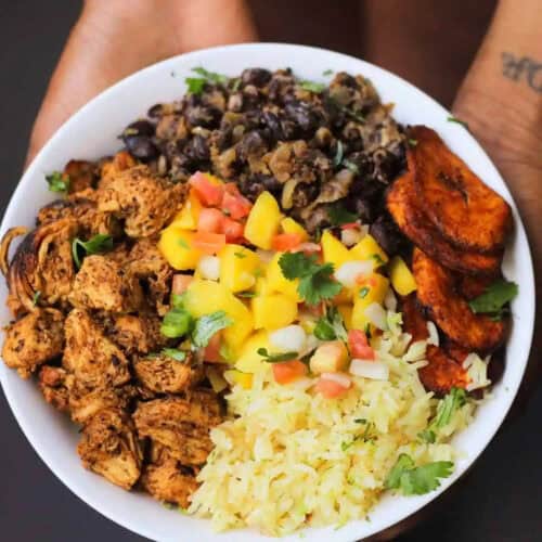 A bowl, brimming with a vibrant assortment of food, features grilled chicken cubes, black beans, fried plantains, diced mango salsa, yellow rice, and fresh cilantro garnishes. The bowl is showcased in two hands with part of a tattoo visible on one wrist—a perfect blend for delicious rice recipes.