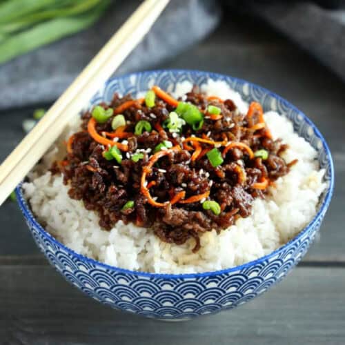 A blue and white patterned bowl filled with white rice, topped with cooked ground meat, shredded carrots, and chopped green onions. A pair of wooden chopsticks rests on the rim of the bowl. The background features a dark surface and some green onions—perfect for Rice Bowl Recipes enthusiasts.