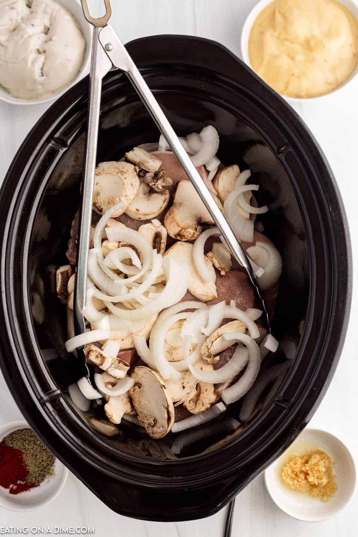 Pork chops in the slow cooker topped with slice mushrooms and onion slices with tongs in slow cooker. Bowls of cream of soups, minced garlic and seasoning on the side