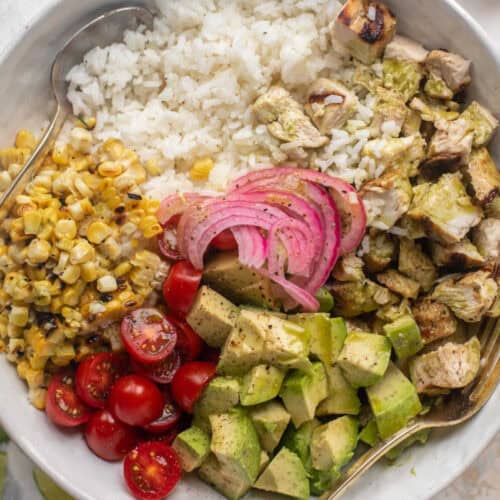 A bowl filled with white rice, grilled chicken pieces, grilled corn kernels, cherry tomatoes, avocado chunks, and pickled red onions. A spoon is placed in the bowl. The ingredients are arranged in a visually appealing, colorful presentation—a perfect addition to your Rice Bowl Recipes collection.