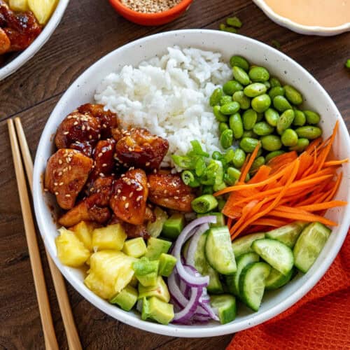 A bowl filled with an assortment of food items including white rice, edamame, sliced carrots, cucumber chunks, red onion slices, diced avocado, pineapple pieces, and sesame seed-topped tofu or chicken in a sauce. Chopsticks are laid beside the bowl on a wooden surface—one of the best Rice Bowl Recipes to try.