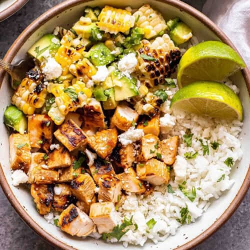 A bowl filled with grilled chicken chunks, charred corn, diced avocado, crumbled cheese, white rice, and cilantro, garnished with two lime halves on the side and drizzled with a dark sauce. Perfect for those exploring new rice bowl recipes.