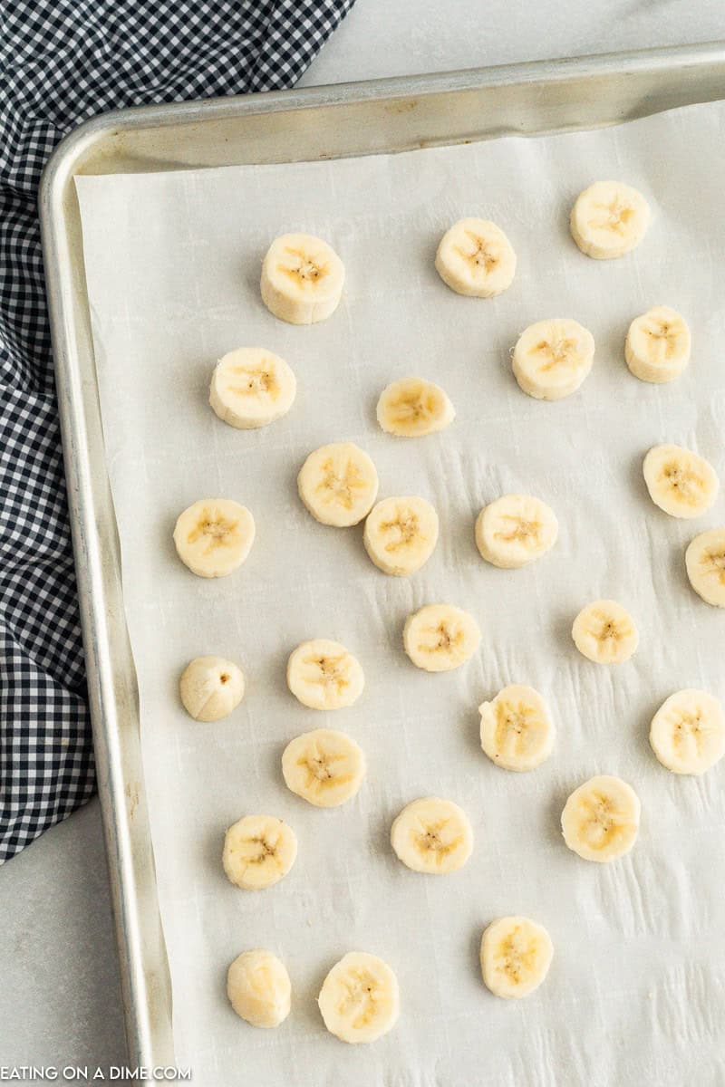 A baking tray lined with parchment paper holds evenly spaced banana slices, illustrating how to freeze bananas effectively. The tray rests on a light-colored surface, partially covered by a black and white checkered cloth.