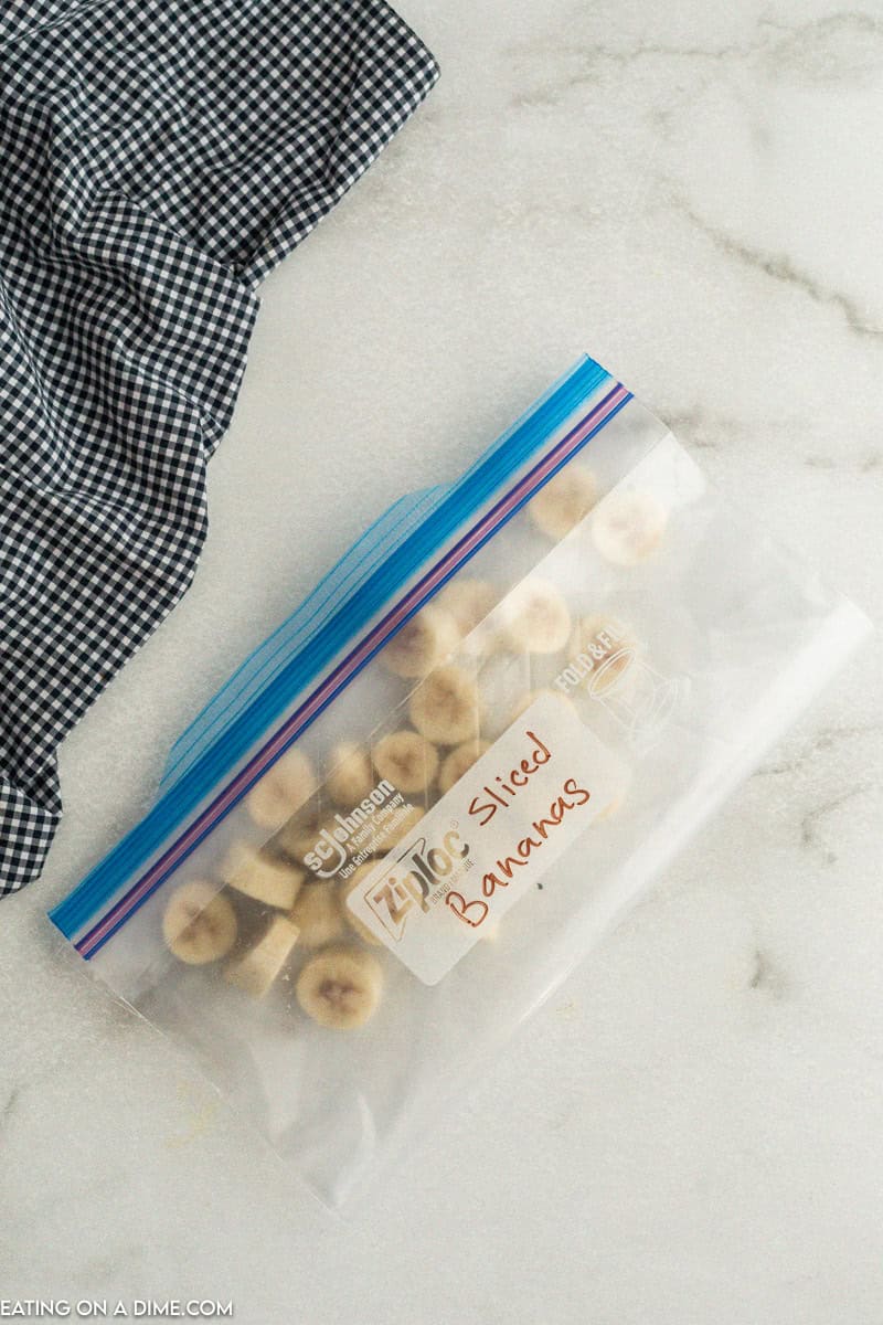 A clear Ziploc bag labeled "sliced bananas" contains small slices of the fruit. Perfect for those wondering how to freeze bananas, the bag is placed on a light-colored marble surface with a black and white checkered cloth partially visible to the side.