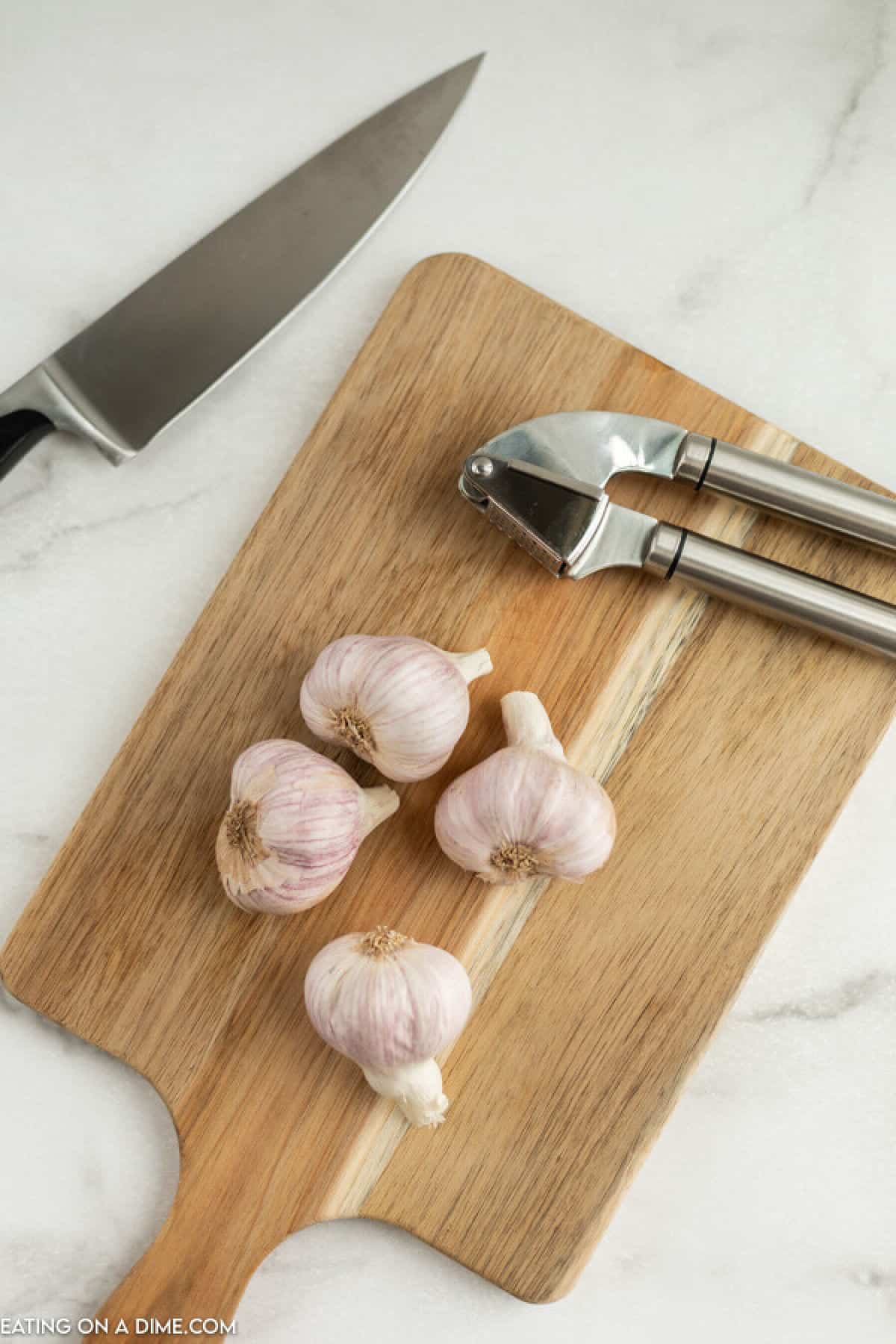 Close up image of items needed to mince garlic - garlic cloves, cutting board, knife and a garlic press. 