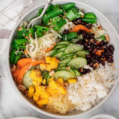 A colorful Buddha bowl filled with fresh vegetables and rice recipes. The bowl includes sliced cucumbers, snap peas, black beans, shredded cabbage, carrot slices, mango chunks, pickled vegetables, peanuts, and a portion of steamed white rice. A fork rests on the edge.