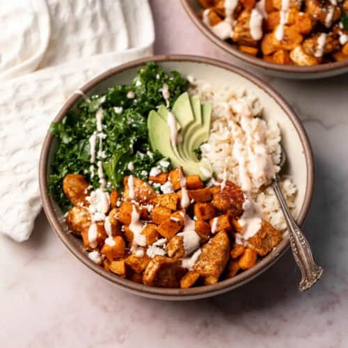 A bowl filled with a balanced meal, featuring cooked chunks of sweet potato, sliced avocado, steamed rice, and kale. Drizzled with a creamy sauce and topped with crumbled feta cheese, this delightful creation is an ideal addition to your rice bowl recipes collection. A spoon is placed inside the bowl.