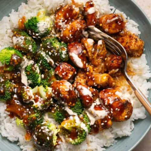 A delectable bowl meal featuring a plate of rice topped with grilled chicken bites, roasted broccoli, and drizzled with teriyaki sauce and creamy dressing. This dish exemplifies one of the best rice recipes, garnished with sesame seeds and accompanied by a metal fork on the side.