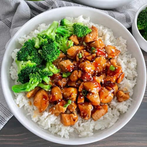 A white bowl filled with perfectly cooked rice topped with cubes of teriyaki chicken, garnished with sesame seeds and chopped green onions, sits on a table. Steamed broccoli florets are placed on one side of the bowl, adding a pop of green to the dish—a delightful addition to any rice bowl recipes collection.