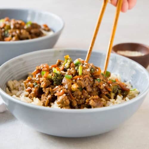 A bowl of rice topped with a flavorful mixture of seasoned beef, chopped vegetables, and garnished with green onions and sesame seeds. A hand is using chopsticks to pick up some of the beef mixture from the bowl. Another similar bowl from popular recipes is visible in the background.