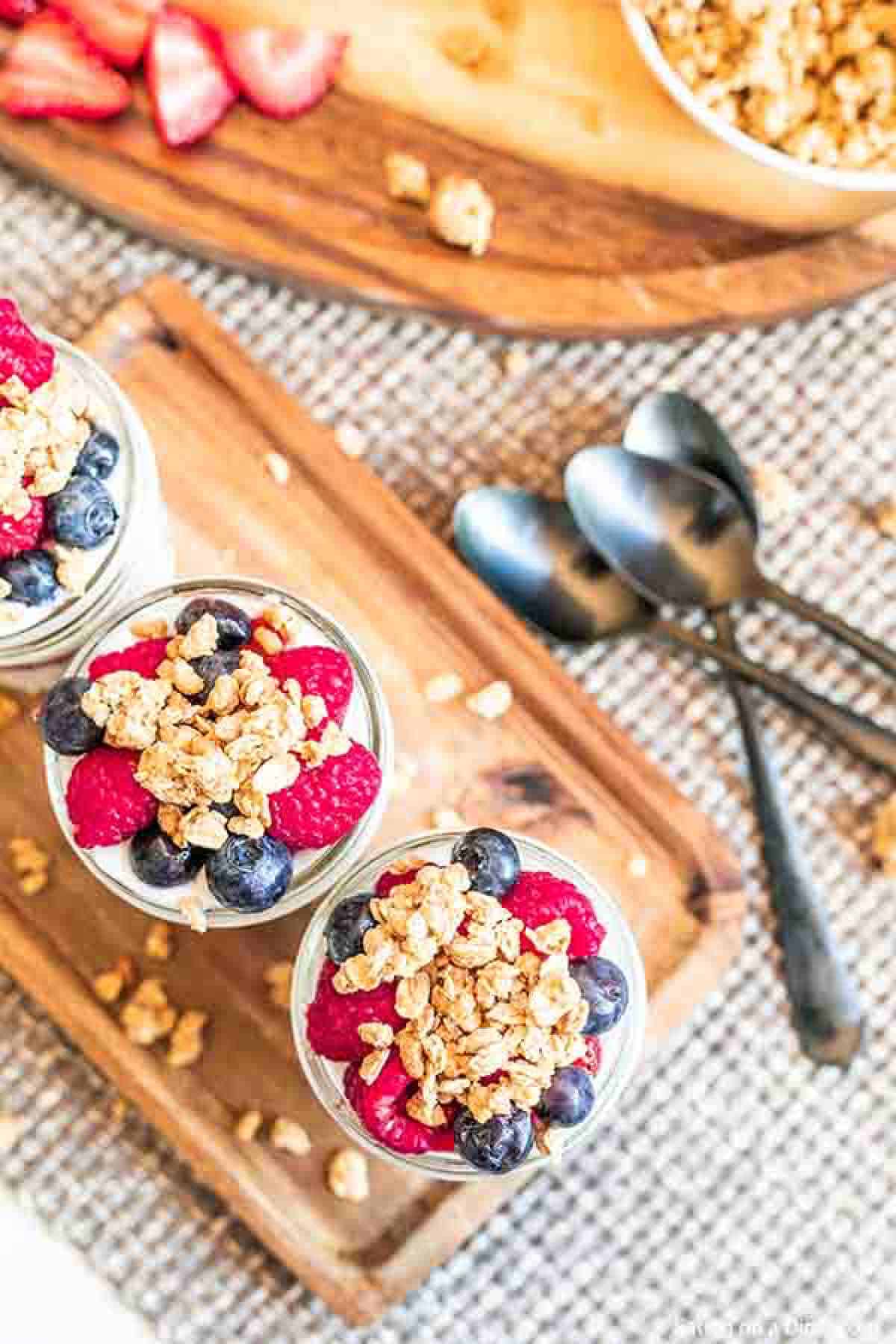 Fruit and yogurt parfaits on a wooden platter topped with granola, raspberries and blueberries