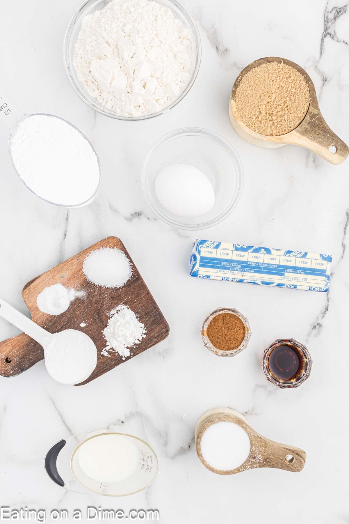 A top-down view of baking ingredients for cookies on a marble countertop. Items include flour, light brown sugar, powdered sugar, a stick of butter, an egg, vanilla extract, ground cinnamon, and other assorted ingredients on wooden boards and measuring spoons. Text reads "Eating on a Dime.com".