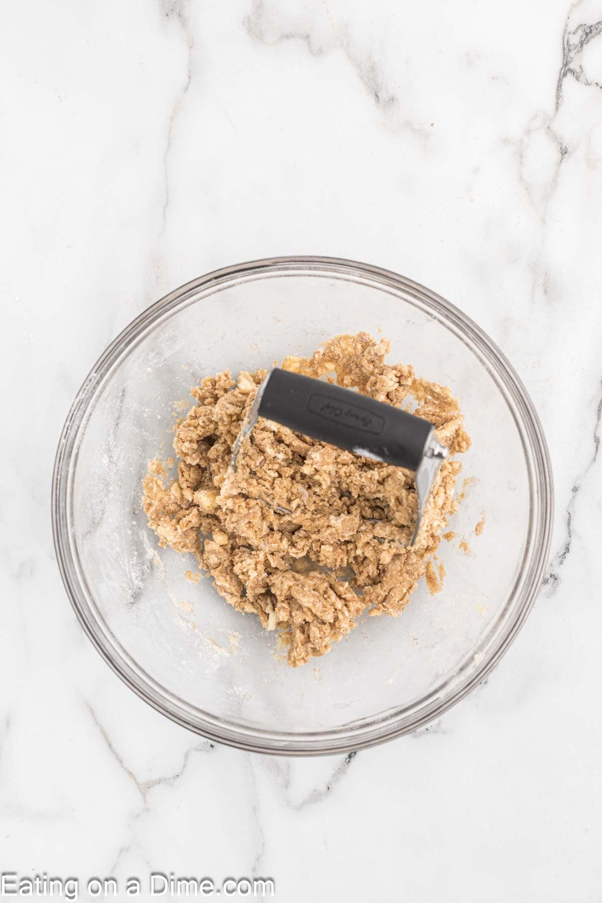 A glass bowl containing a crumbly mixture sits on a white marble surface. A black-handled pastry cutter is resting in the bowl, partially embedded in the mixture. The text at the bottom reads "Eating on a Dime.com". Perfect for baking cookies or cake!