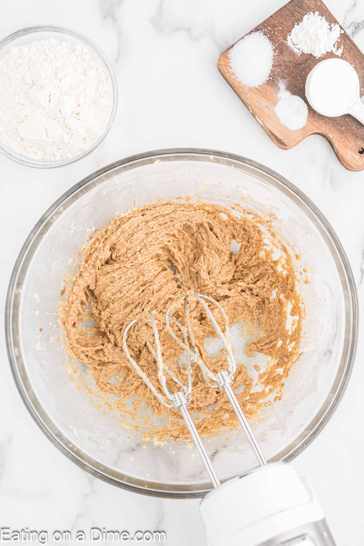 A glass mixing bowl with brown cookie dough and a hand mixer inside. Nearby, there is a small bowl of flour and a wooden board holding salt, baking soda, and a measuring spoon with white powder—all set on a marble surface, ready to create delicious cookies.