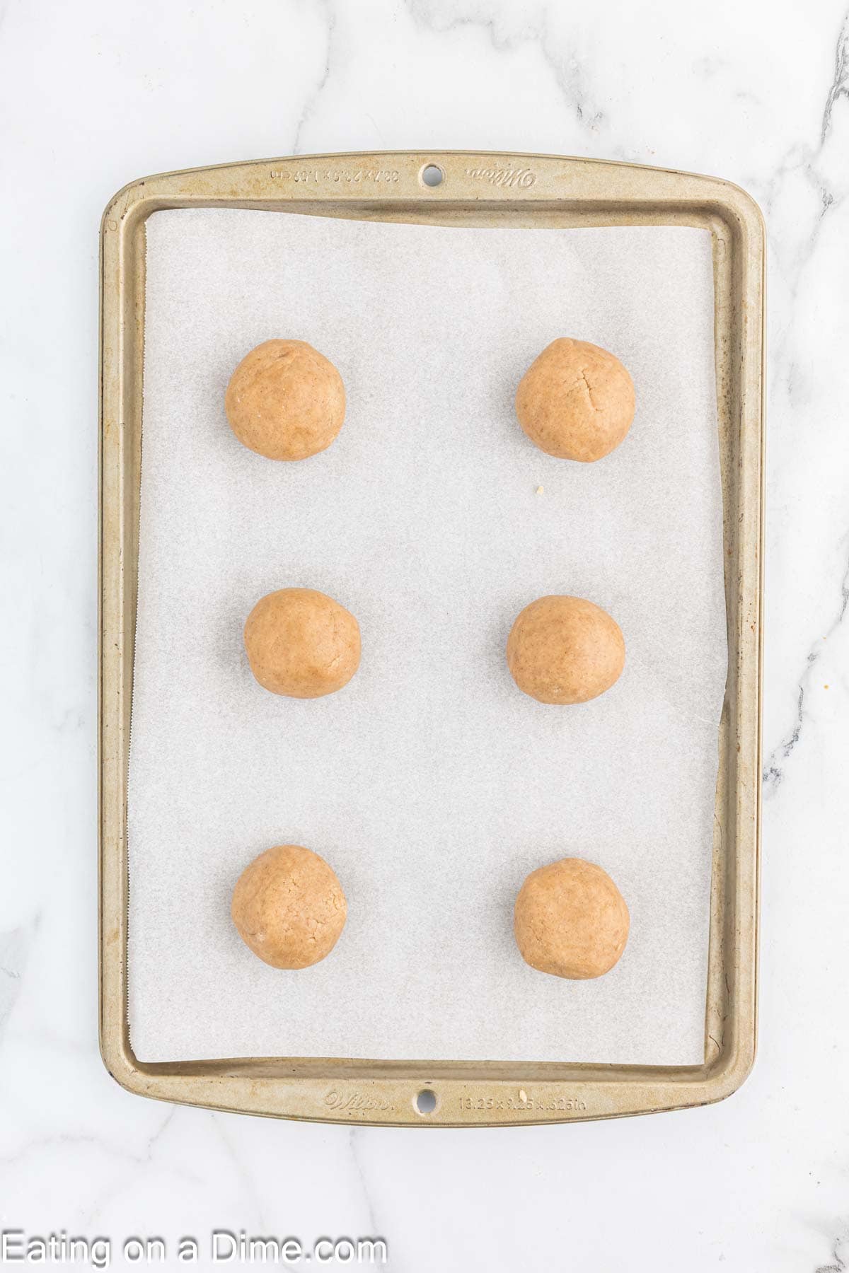 A baking sheet lined with parchment paper holds six evenly spaced, unbaked cookie dough balls, perfect for your next coffee break. The text "Eating on a Dime.com" is visible at the bottom left corner of the image. The background is a white marble surface.